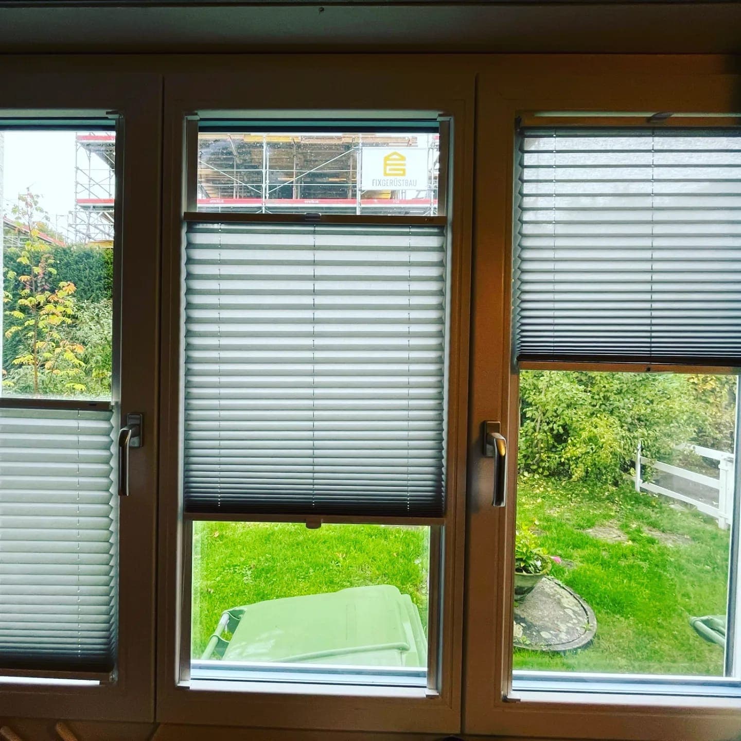 Hellgraue Plissees an drei Fensterflügeln eines Wohnraums, teilweise geöffnet mit Blick in den Garten